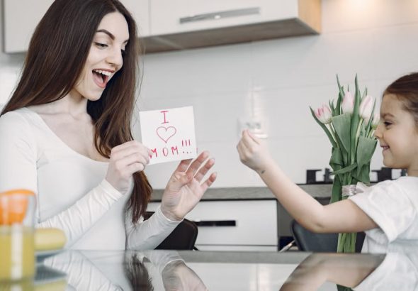 A mother and daughter sharing a joyful moment with flowers and a card in a kitchen.