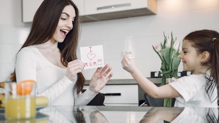 A mother and daughter sharing a joyful moment with flowers and a card in a kitchen.